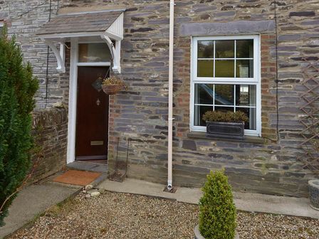 Stone exterior wall with a dark wooden door under a small white roof and a white framed window with a planter box in front of the window at 12 Victoria Terrace Nantlle