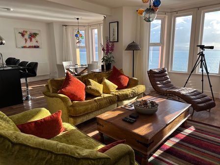A living room with green sofas with red and yellow cushions a wooden coffee table a brown chaise lounge a telescope dining table with white chairs and a kitchen area at Vista Marina - Penthouse in Criccieth