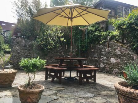 A circular wooden picnic table with benches and a large yellow umbrella in a stone patio garden at 3 Top View Cottages in Salcombe