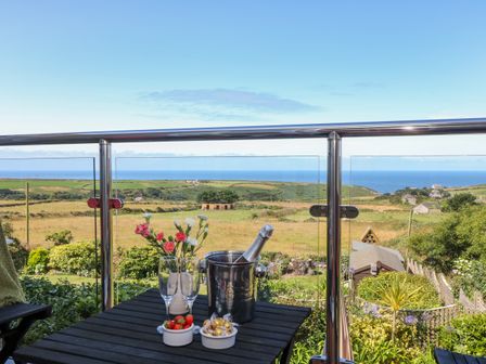 A balcony with a table holding flowers glasses a bucket with a bottle and bowls of strawberries and chocolates overlooking fields and the sea at Sunset in St Just