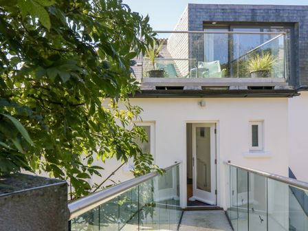 An outdoor walkway with glass and metal railings leading to a white door on a building with an upper balcony with plants and chairs at The Upper Deck in Porthcurno