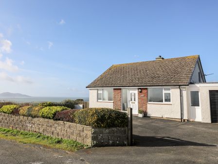 A single-story house with a tiled roof and white walls near the sea with a stone wall and shrubs in front at Yr Orsedd in Llanfaethlu