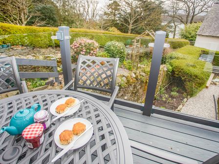 An outdoor patio table with scones jam and a teapot on it on a balcony overlooking a garden at Bryn Teg Cottage in Menai Bridge