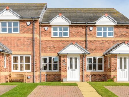 A brick townhouse with white doors and windows a wooden bench and grass lawn at 39 Longbeach Drive in Beadnell