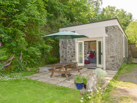 A stone cottage with open double doors a wooden picnic table with umbrella on the patio and plants around at Engine House in Bridford