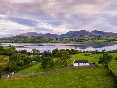 A rural landscape with a small white house near a lake and green fields with mountains in the background at Lough Island Reavy Cottage Kilcoo Mourne Mountains Newry County Down