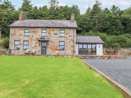 A stone house with a dark door and windows surrounded by a lawn and trees at The Cottage in Llaniestyn