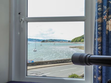 A window view showing a coastal road with boats in the water and trees in the background at Gadlys House - Waters Edge in Beaumaris