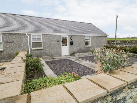 A stone wall and garden with shrubs in front of a grey pebble dash house with a tiled roof at Ysgubor Llwyd in Valley