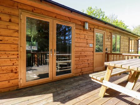 Wooden cabin exterior with double glass doors a side door and a wooden picnic table on a wooden deck at Lodge Three Brighton near Summercourt