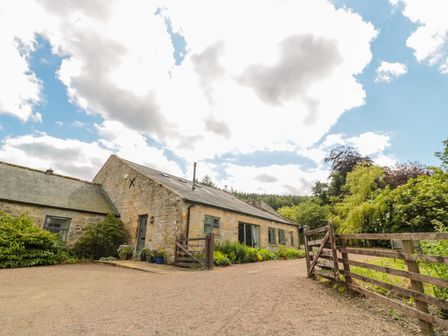 An exterior view of a stone house with a gravel driveway and wooden gate surrounded by bushes and trees at The Steadings in Rothbury