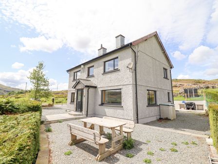 An outdoor gravel area with a wooden picnic table and benches next to a two-story gray house with black window frames at Craig Yr Ronwy Bala