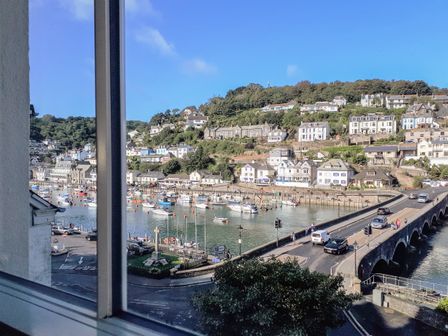 A view of a harbor with boats docked near a bridge and houses on a hillside seen through a window at Harbour View Apartment in Looe