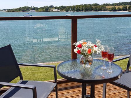 A glass table with flowers and two glasses of red drink between two chairs on a wooden deck overlooking water at Delfryd in Y Felinheli