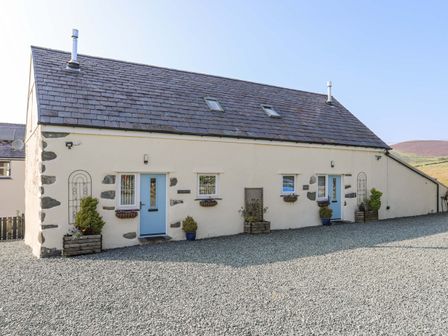 A cream colored cottage with blue doors and small windows in a gravel yard at Ffynnon Bach in Deiniolen
