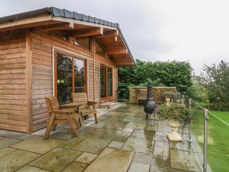 A wooden cabin with glass doors a stone patio wooden chairs a chiminea glass railing and flower pots at Nicky Nook Lodge in Scorton