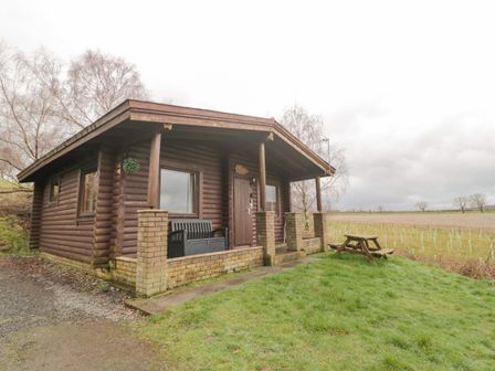 A small log cabin with a covered porch and bench next to a grassy area with picnic table at Glanton in Longframlington