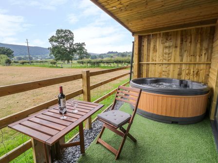 A balcony with a wooden table and chair and a hot tub overlooking a field at Cilan Lodge in Llandrillo