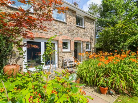 A stone house exterior with white framed windows and doors surrounded by orange flowers and green plants with garden chairs on the patio at Coth Skyber in Hayle