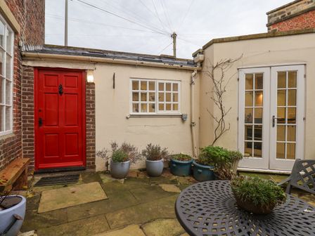 A small courtyard with a red door, potted plants, a metal table with a planter, and white French doors at 20A St Hildas Whitby