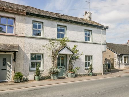 A street view of a white house with green door and window frames with plants outside at 75 Station Road in Cark In Cartmel