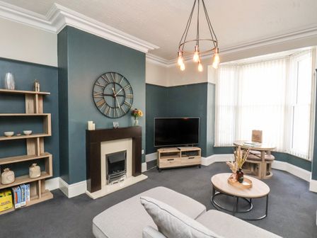 A living room with a fireplace a wall clock a television on a stand a round coffee table and a dining table near a bay window at Church View in Whitby