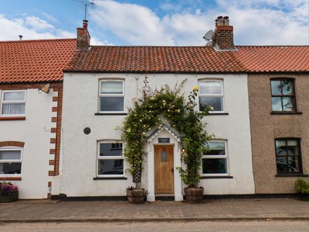 A street view of a terraced house with a wooden door framed by flowering plants at Burton Fleming near Hunmanby