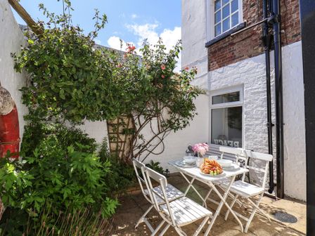 A small outdoor patio with a white table and chairs set with breakfast items and surrounded by plants and brick walls at Albany Cottage in Whitby