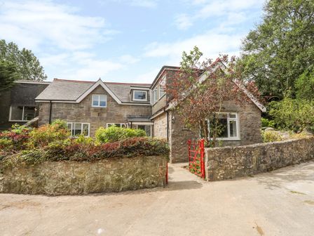 A stone house with a red metal gate and a stone wall surrounded by bushes and trees at Gorwel in Moelfre
