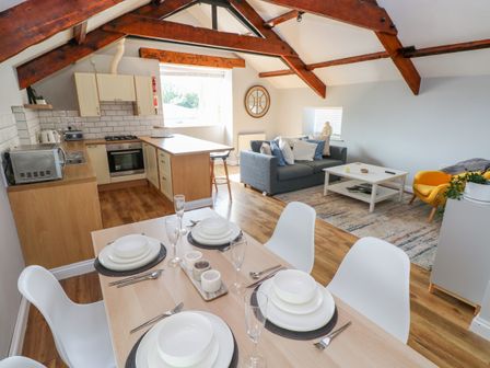 An open plan kitchen and living area with a dining table set with white plates and cutlery a gray sofa a yellow chair and wooden ceiling beams at The Old Brewery in Saundersfoot