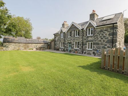 A stone house with multiple windows a blue door a wooden bench and a lawn with a stone outbuilding and picnic table at Tyn Ffynnon in Dyffryn Ardudwy