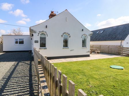 The exterior of a white house with three arched windows a chimney a wooden fence a gravel driveway and a grassy yard with a patio and picnic table at Senafe in Widemouth Bay