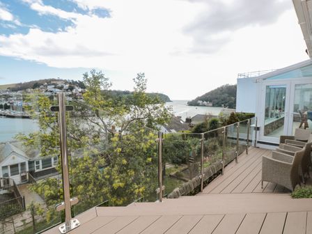 An outdoor patio with wicker chairs and glass railing overlooking a river and hillside at Estuary View House and Annexe in Dartmouth