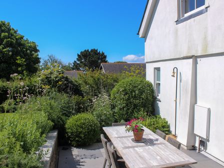 A patio with a wooden dining table and chairs next to a white house with a garden surrounding it at Willow Cottage in Dartmouth