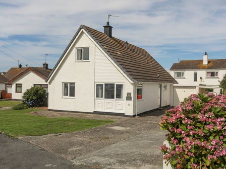 A white detached house with a triangular roof and a driveway with pink flowers in front at Crows Nest Rhosneigr