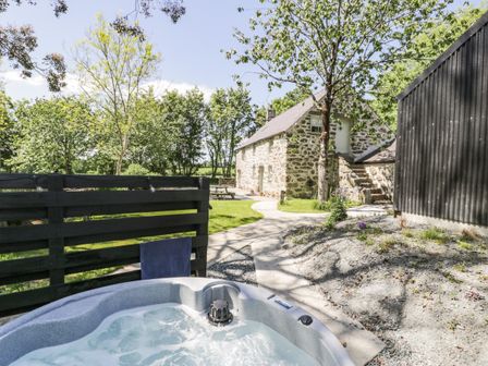 An outdoor hot tub near a stone house surrounded by trees and a wooden fence at Stabla in Chwilog
