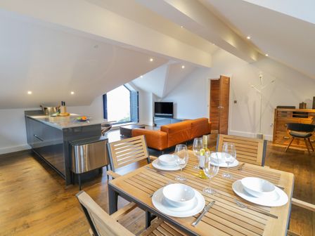An open-plan living space with a wooden dining table set for four next to a kitchen island and a seating area with a brown sofa and television at Lake View in Bowness-On-Windermere