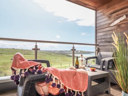 A balcony with black wicker chairs a red blanket a bottle and glasses overlooking a grassy field and ocean at 8 Cribbar in Newquay