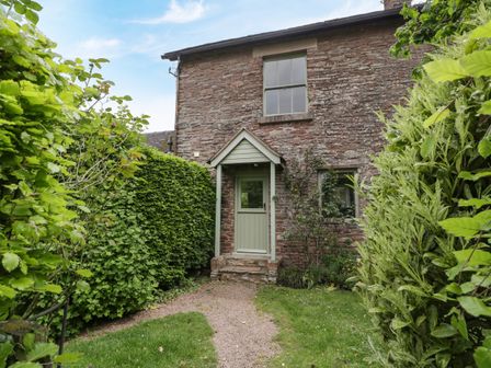 Front view of a stone cottage with a green door and a small porch surrounded by hedges and plants at Woodlands Cottage in Docklow near Leominster