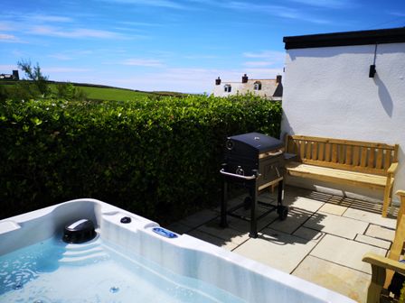 An outdoor patio with a hot tub a barbecue grill and wooden benches surrounded by greenery at Trevena Lodge in Tintagel