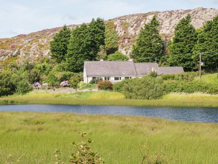 A house behind trees near a pond with rocky hills in the background at Glan Gors in Llanerchymedd