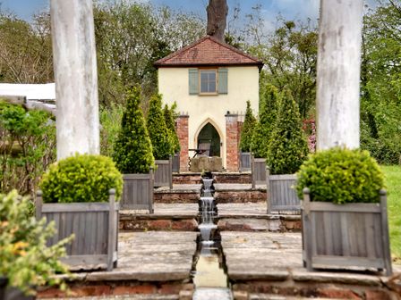 A garden with a small building at the end of a stone pathway with water features and potted plants at The Folly in Weobley