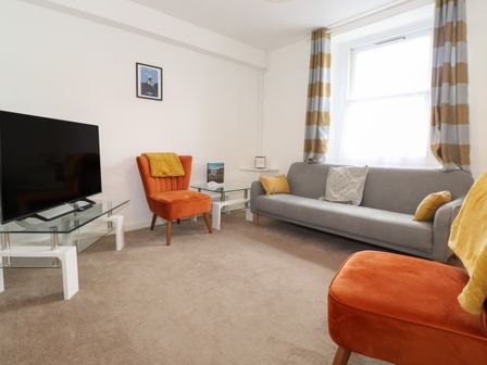 A living room with a large window with striped curtains a gray sofa with cushions two orange chairs a television on a glass stand and a small glass side table at Holly Cottage in Keswick