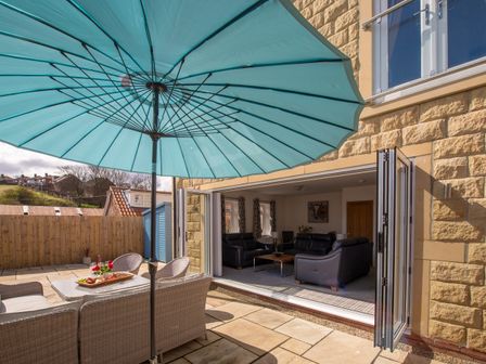 An outdoor patio with a large blue umbrella a wicker seating set and a table with food next to a living room with black leather sofas at Lemonade Cottage in Whitby