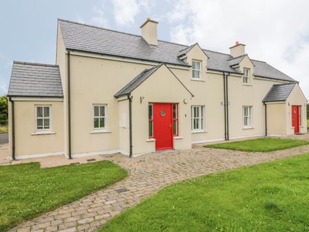 A beige two story building with red doors and a paved path surrounded by grass at No 6 An Seanachai Holiday Homes Ring nr Dungarvan County Waterford