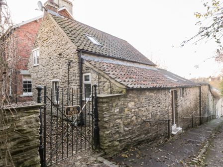 A stone house with a tiled roof behind a black metal gate labeled Hill House on a sloped stone pathway at Dovecote in Pickering