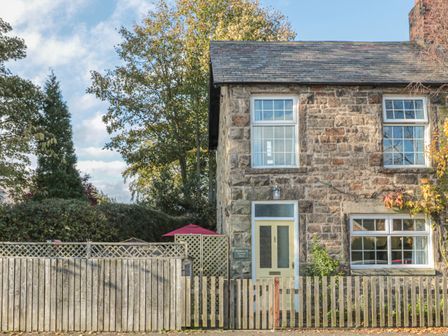 A stone house with a yellow door and three windows behind a wooden fence at Wordsworth Cottage West in Longframlington