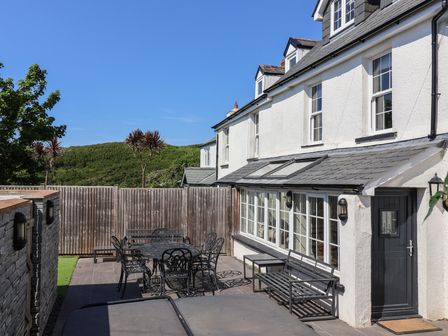 An outdoor patio area with a metal table and chairs a bench and a white house with large windows at Rosebank Cottage in Mortehoe