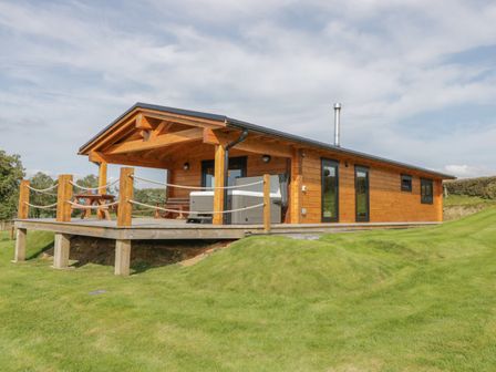 An exterior view of a wooden log cabin with a covered porch and picnic table on a grassy lawn at Bryn Eiddon Log Cabin in Machynlleth