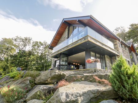 A modern two-story house with glass balcony and stone exterior surrounded by rocks and trees at Lakeside at Louper Weir in Bowness-On-Windermere